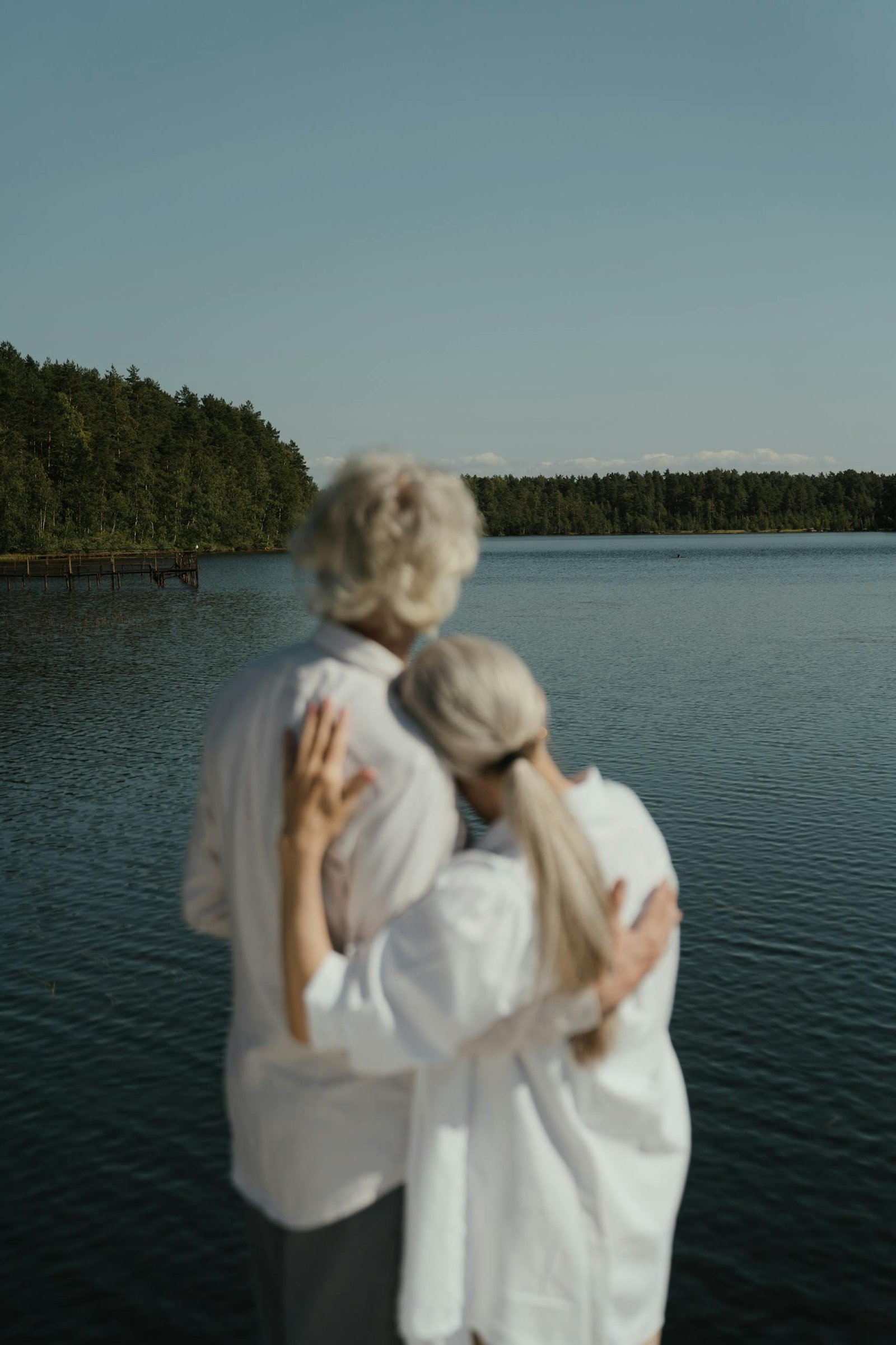 A senior couple shares a tender embrace beside a calm, scenic lake under clear skies.