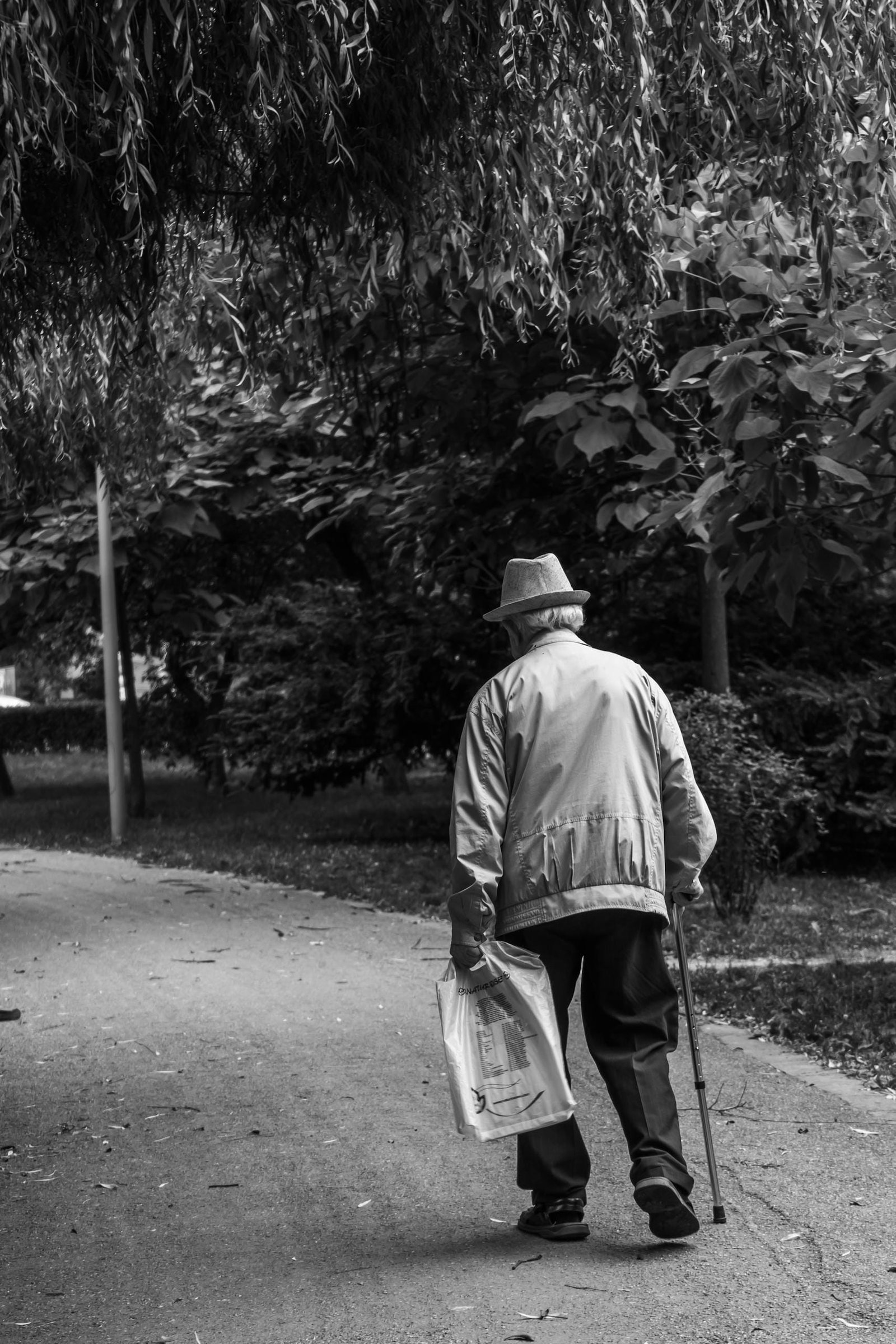 Elderly man in a hat walking on a park path with a cane, captured in monochrome.