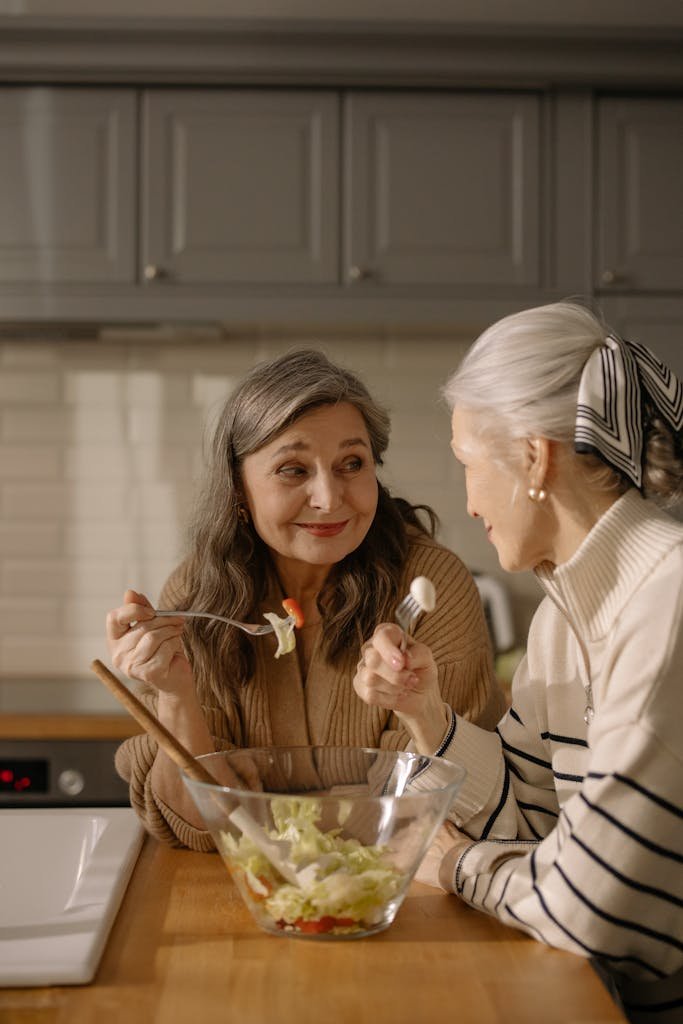 Two senior women share a light-hearted conversation over a fresh salad in a cozy kitchen.