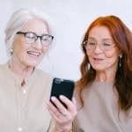 Two senior women sharing a moment of laughter while using a smartphone indoors.