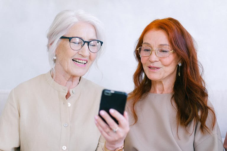 Two senior women sharing a moment of laughter while using a smartphone indoors.